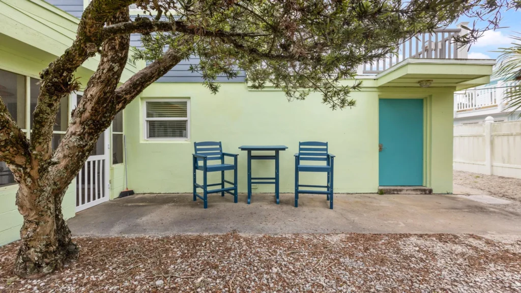 Shaded outdoor seating area with blue chairs and table on concrete patio beneath a large tree.