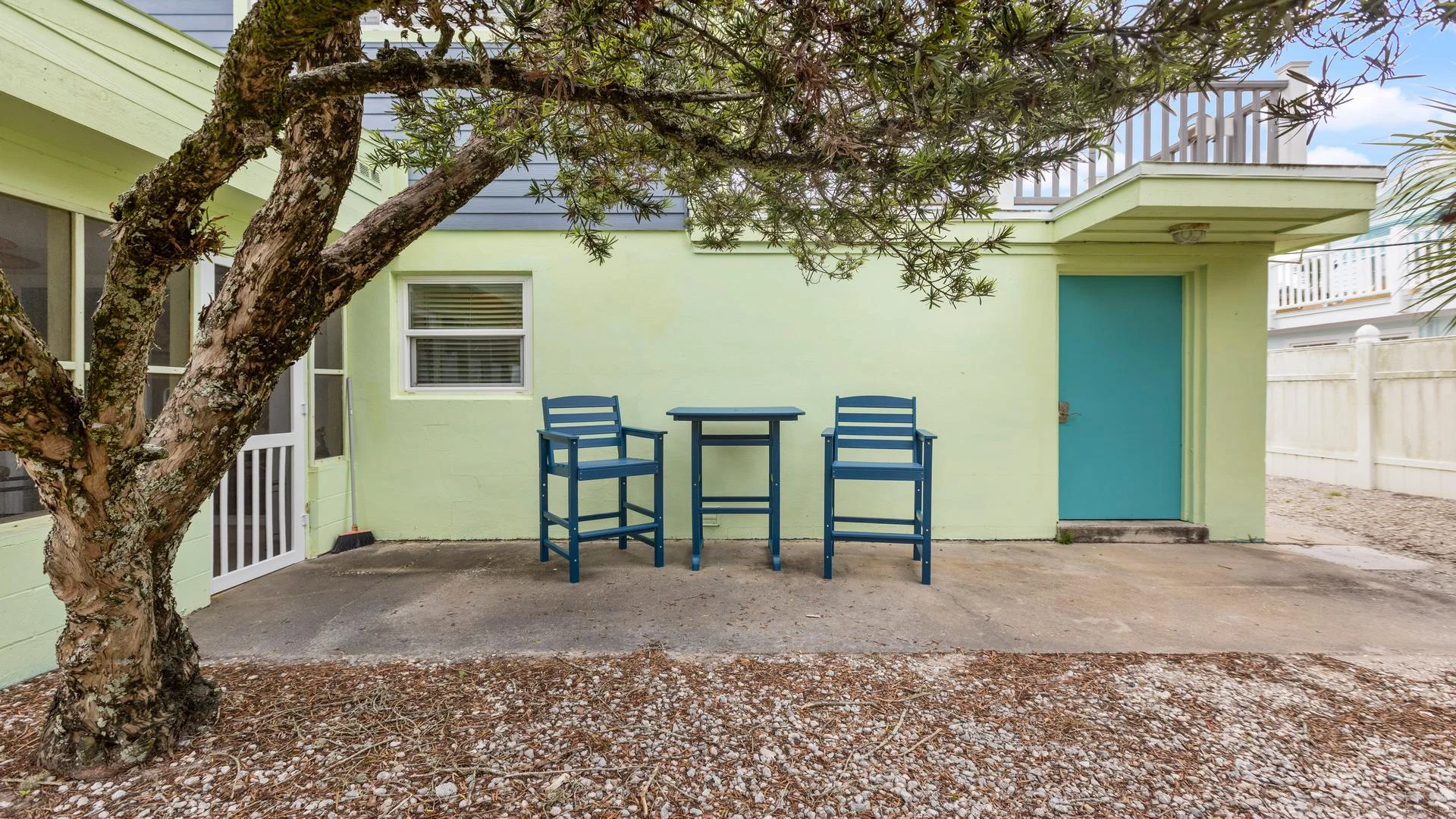 Shaded outdoor seating area with blue chairs and table on concrete patio beneath a large tree.