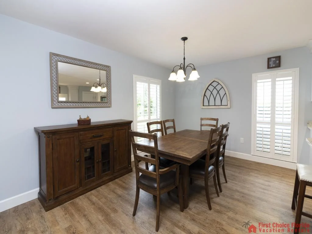 Dark wood dining table with six chairs sits beneath a five-light chandelier by windows with white plantation shutters.