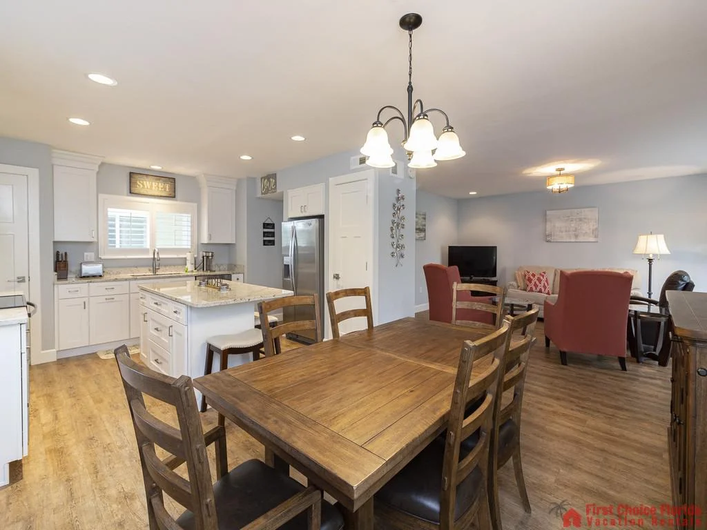 Wood dining table seats six beneath a wrought-iron chandelier, adjacent to a white kitchen island with granite countertop.