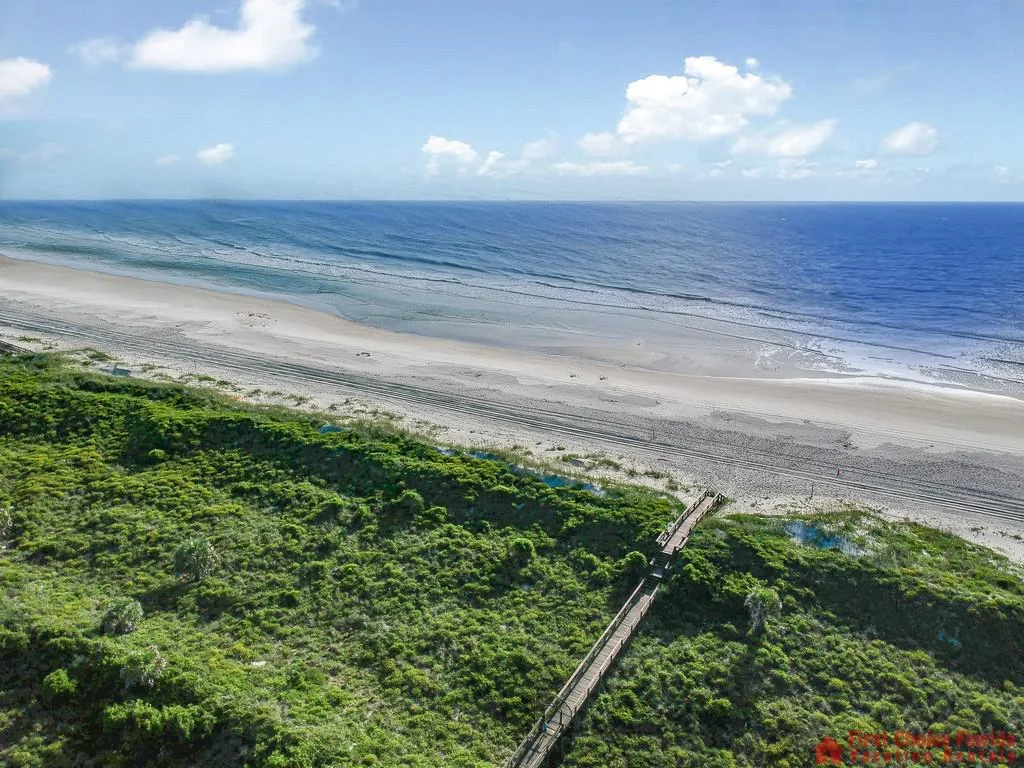 Boardwalk crosses dense green dunes leading to wide sandy beach and blue ocean under a partly cloudy sky.