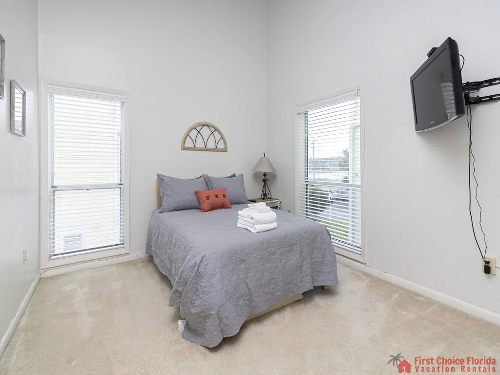 Bedroom with gray bedding, coral accent pillow, beige carpeting, two tall windows, and wall-mounted TV.