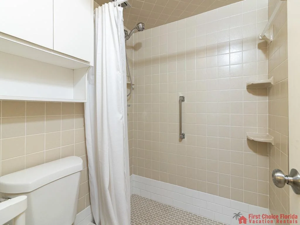 Bathroom shower with beige square tiles, built-in corner shelves, white curtain, and metal grab bar.