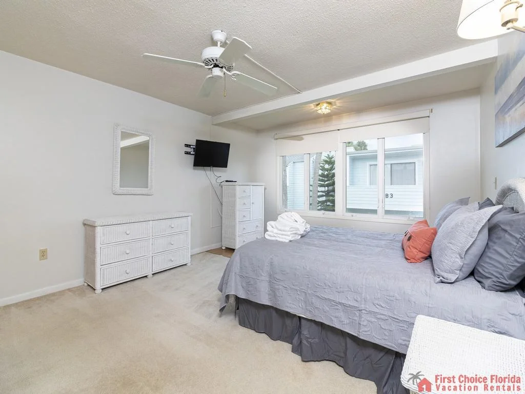 Queen bedroom with gray bedding, white wicker dressers, wall-mounted TV, and large window overlooking courtyard.