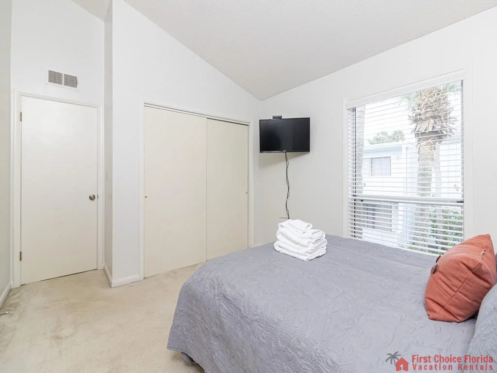 Queen bed with gray quilt and coral pillow faces wall-mounted TV next to window showing palm tree outside.