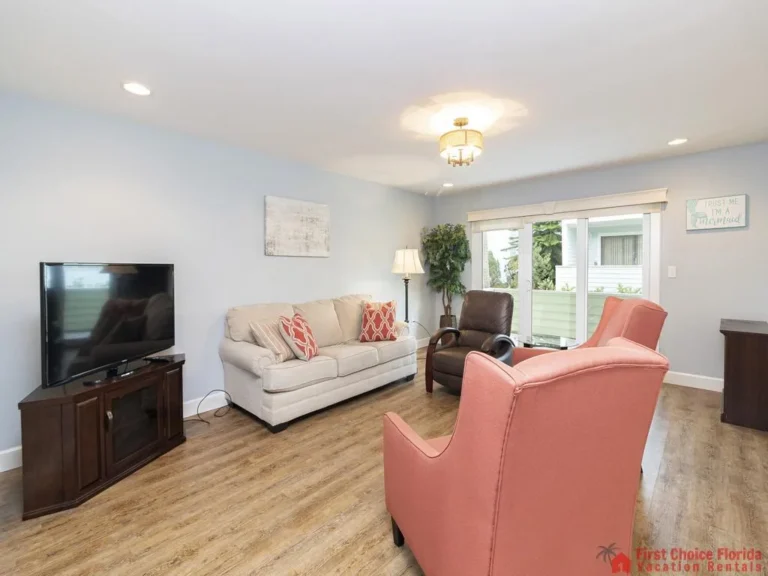 Light wood flooring extends beneath a beige sofa and two coral armchairs facing a corner TV stand.