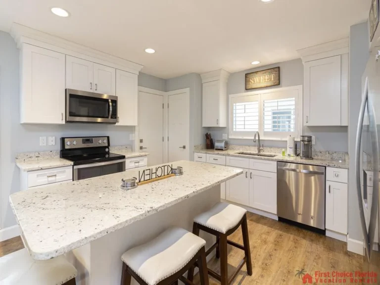 Granite kitchen island with three cushioned stools faces stainless steel appliances and white cabinetry.