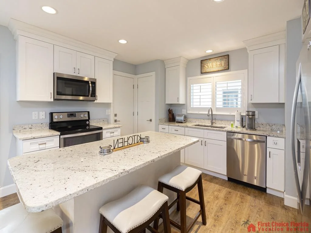Granite kitchen island with three cushioned stools faces stainless steel appliances and white cabinetry.
