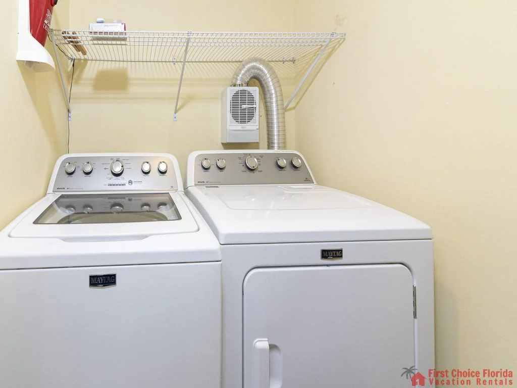 Laundry room with white Maytag washer and dryer beneath a white wire shelf and soft yellow walls.