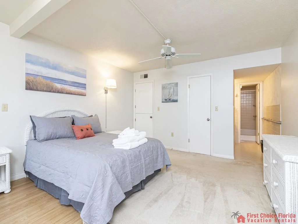 Soft carpeting contrasts with white wicker furniture and coastal-themed artwork in the spacious queen bedroom.