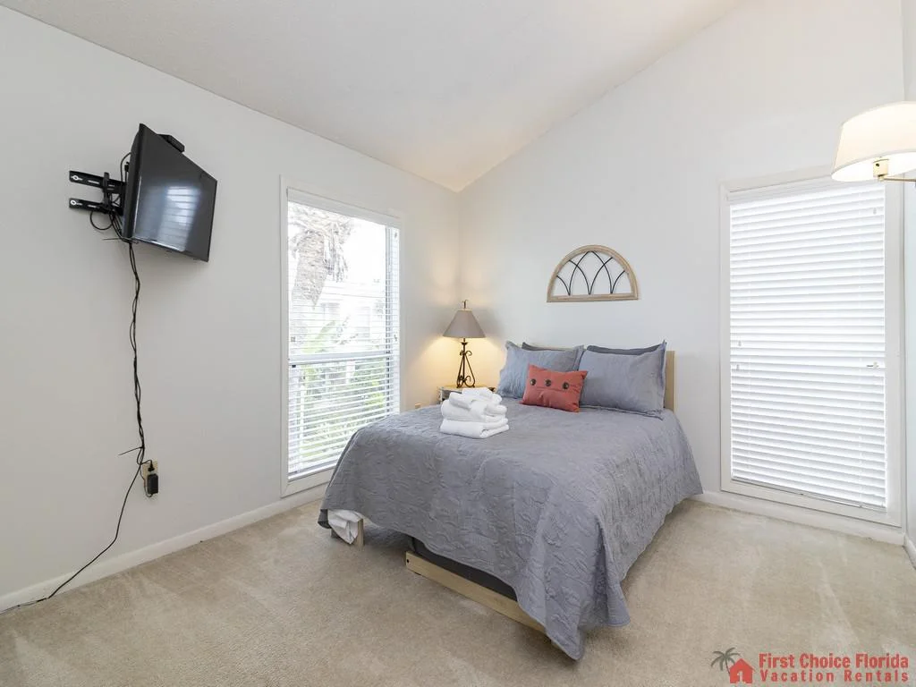 Queen bed with gray bedding and coral accent pillow beneath vaulted ceilings and twin windows with white blinds.