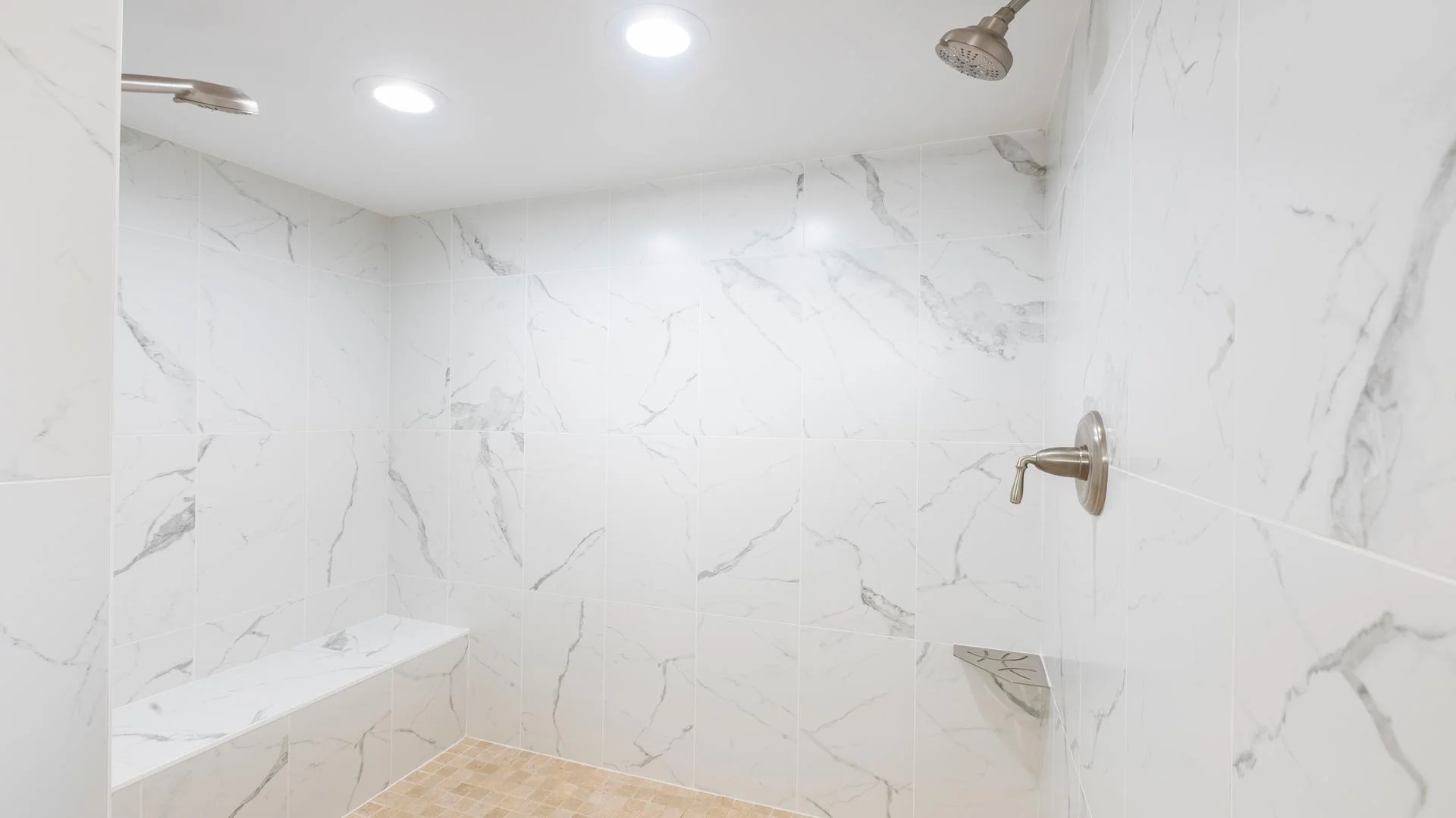 Walk-in shower with white marble-patterned tiles, built-in bench, and brushed nickel fixtures under recessed lighting.