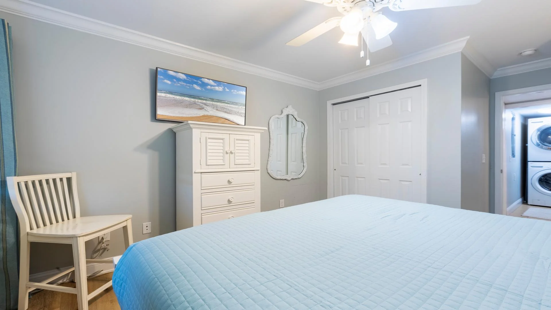 Bedroom with light blue quilted bedspread, white wooden dresser, wall-mounted TV, and stacked washer-dryer visible.