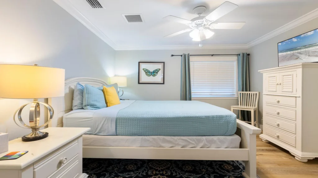 Bedroom with white wooden bed frame, light blue quilt, butterfly art, and dresser under ceiling fan light.