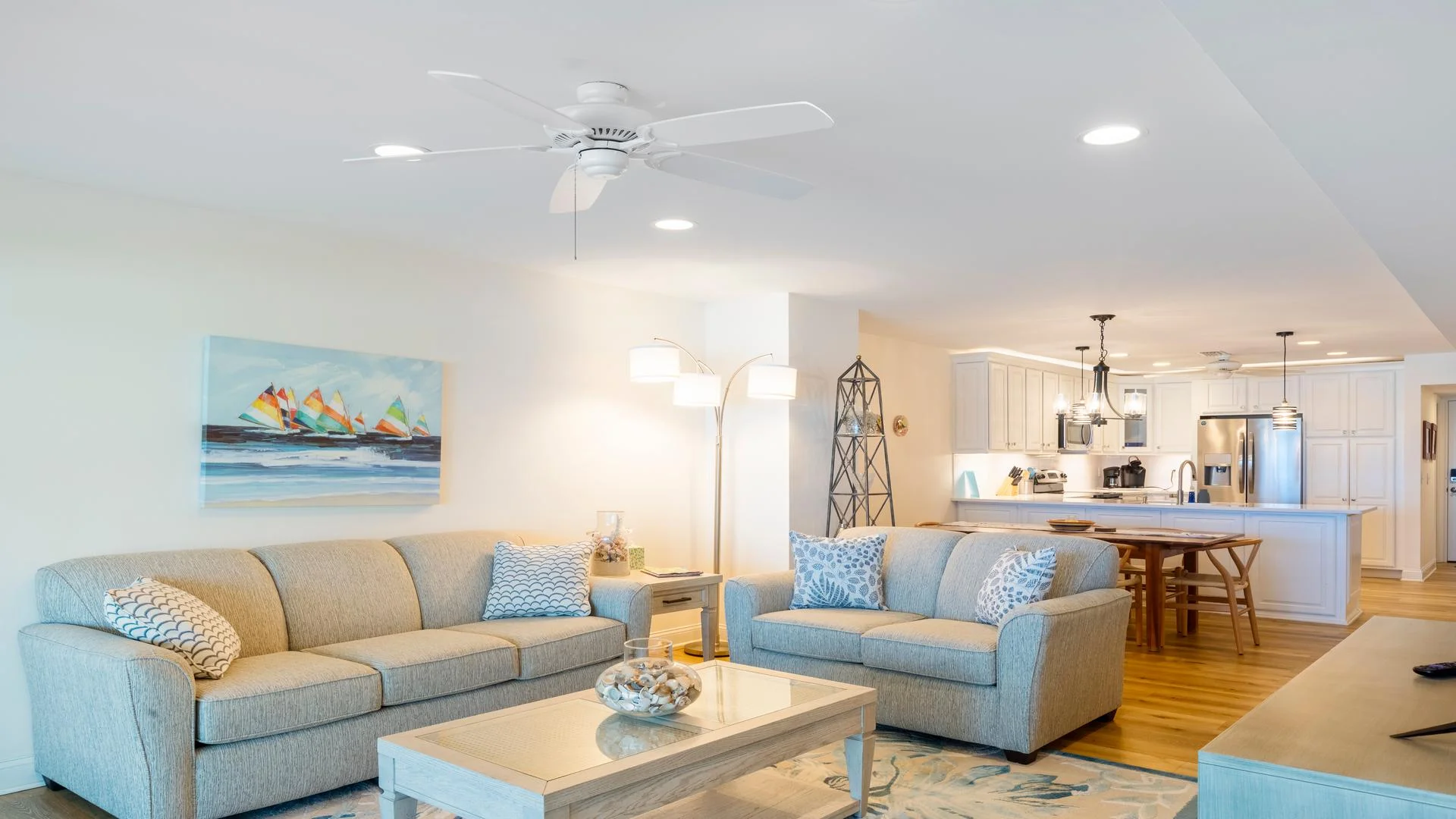 Open-concept living room with beige sofas, patterned throw pillows, coastal artwork, and hardwood floors.