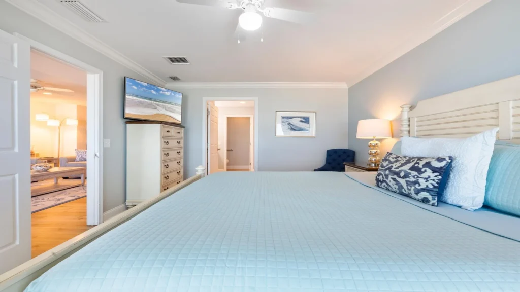 Primary bedroom with light blue quilted bedding, white wood headboard, navy accent chair, and wall-mounted TV.