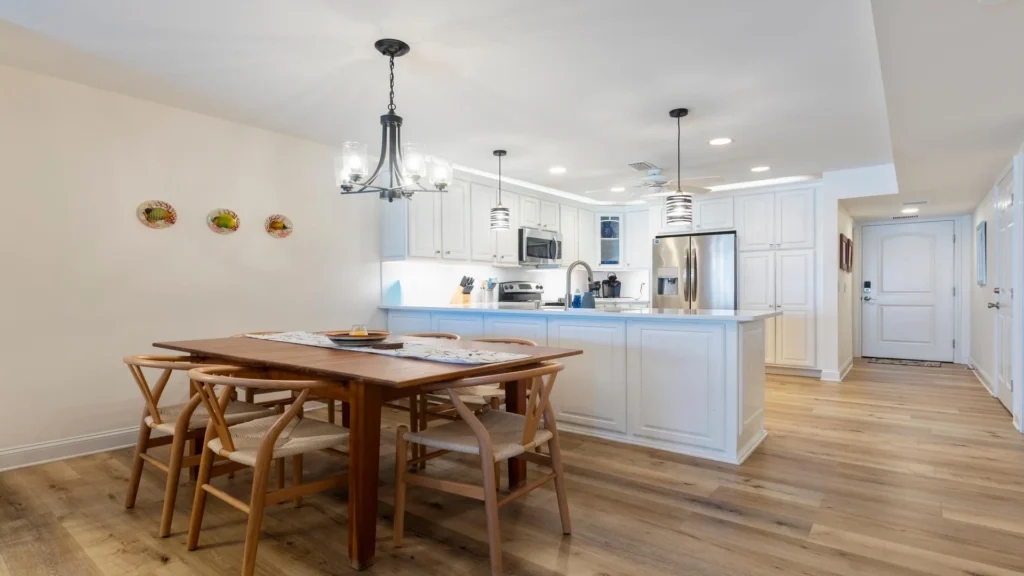 Open-concept dining area with a wooden table and woven chairs beside a white kitchen featuring stainless steel appliances.