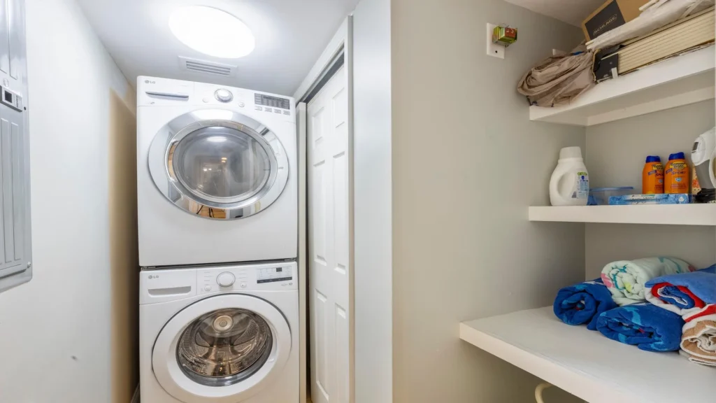 Stacked white front-loading washer and dryer sit under a round ceiling light beside shelves with folded towels and laundry supplies.