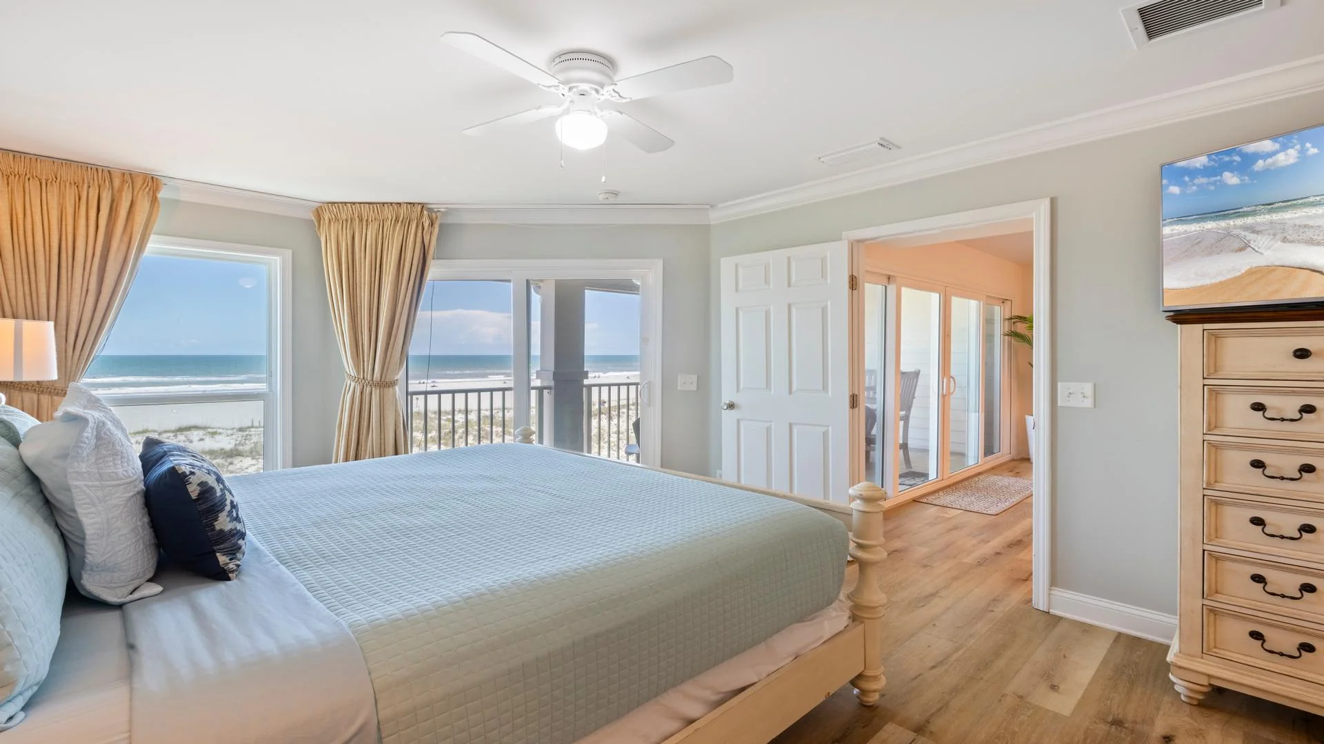 The primary bedroom, with a king bed and a pale blue quilt, faces ocean views through floor-to-ceiling windows and a sliding glass door.