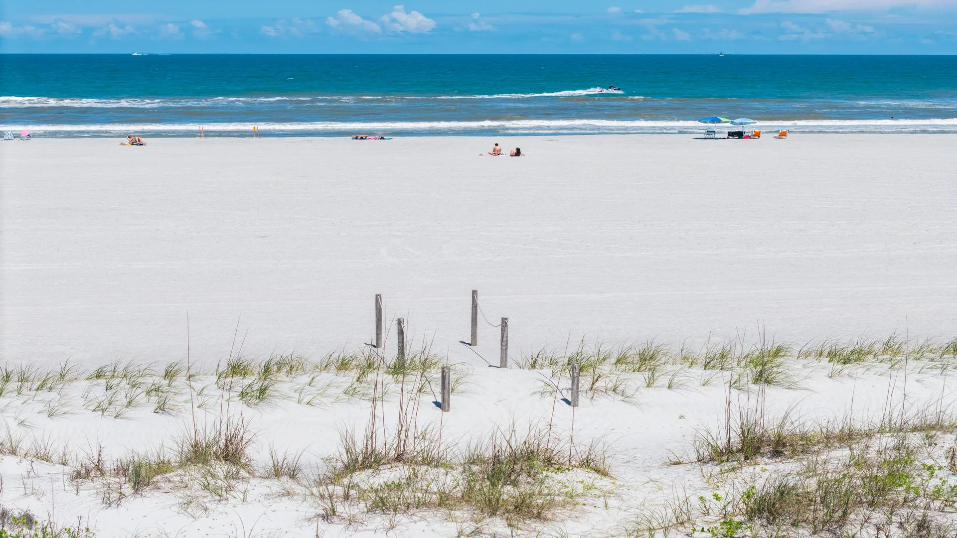 Sandy beach with sparse dune grass and wooden posts leading toward the blue Atlantic Ocean horizon.