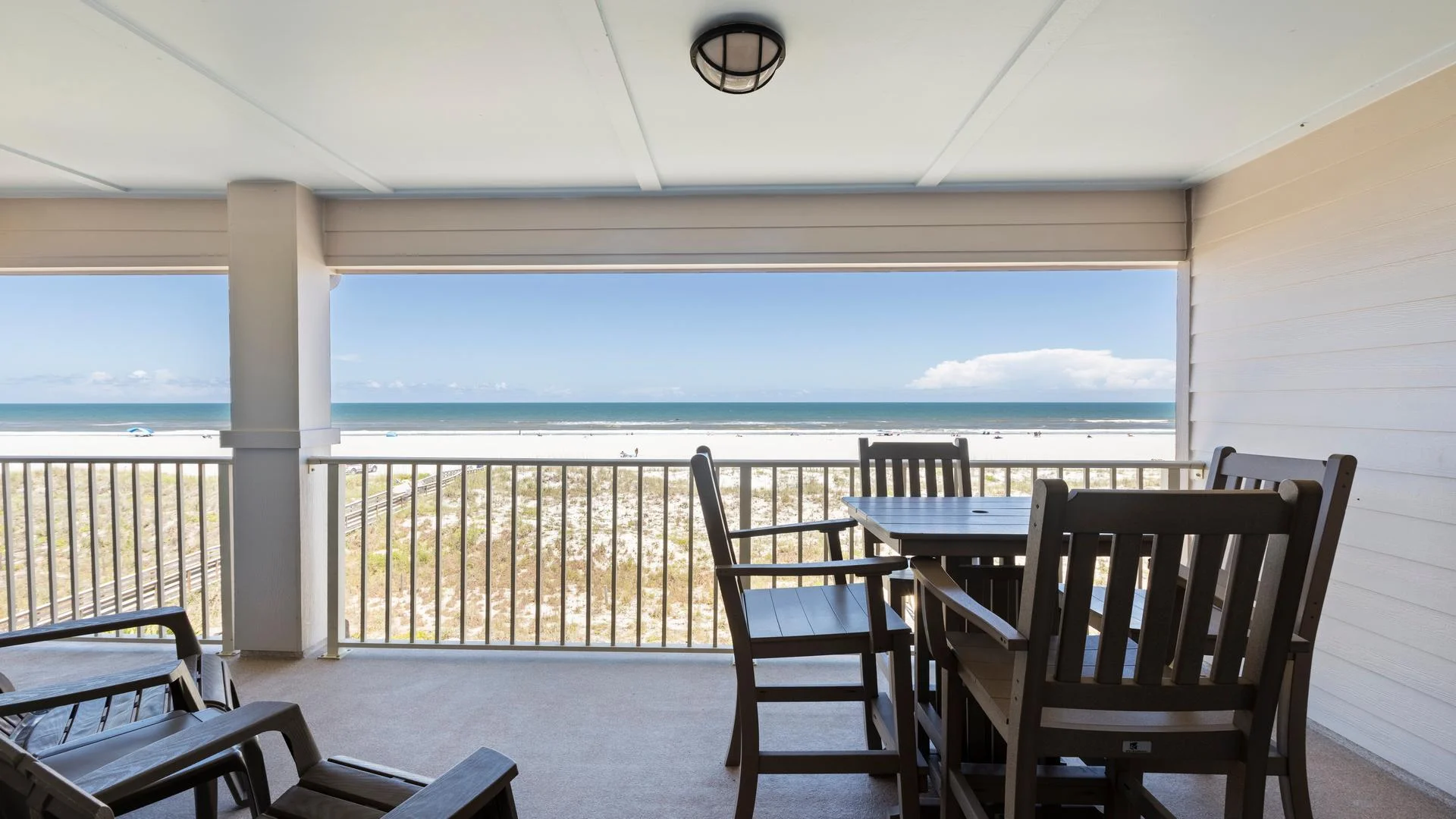 Covered balcony with dark wood dining set and lounge chairs overlooking sandy dunes and the Atlantic Ocean.