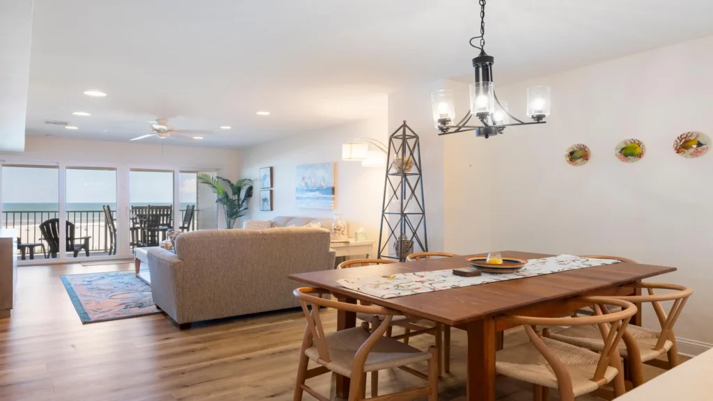 Wood dining table with six woven chairs under a black metal chandelier near the ocean-view living room.