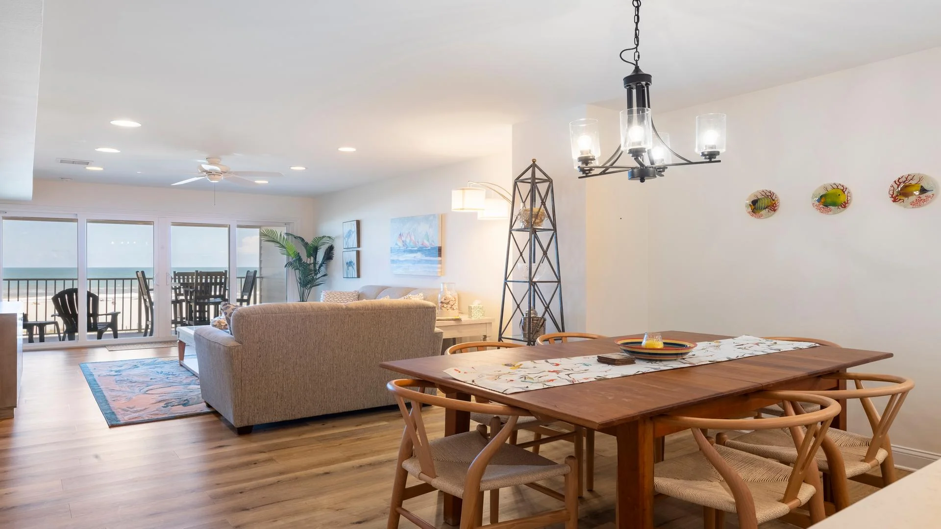 Wood dining table with six woven chairs under a black metal chandelier near the ocean-view living room.
