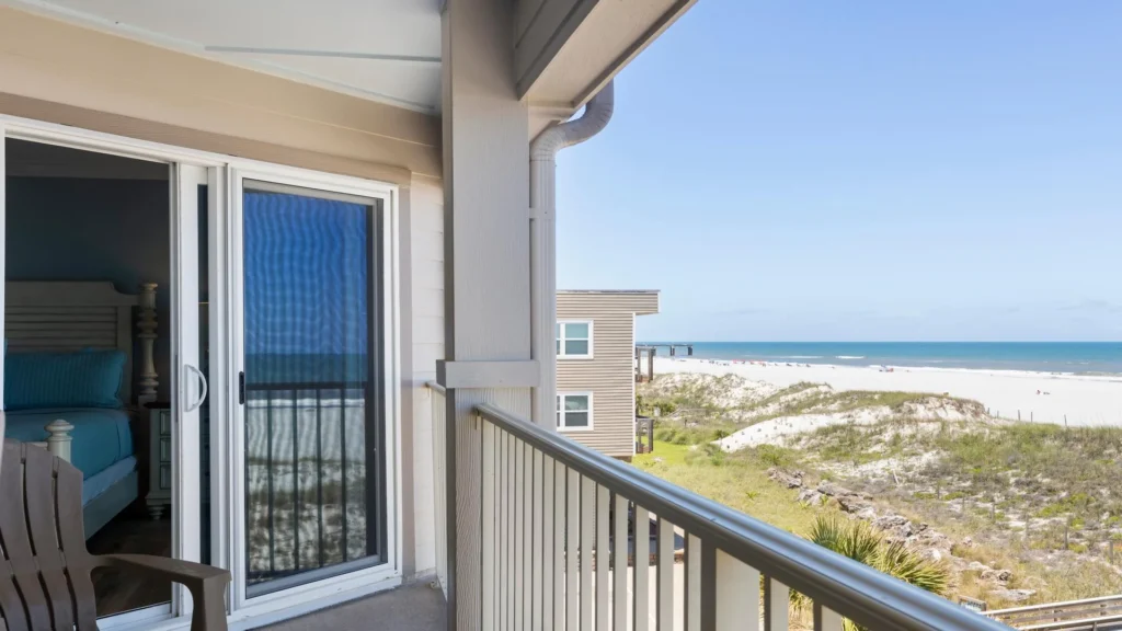Balcony with metal railing overlooks sandy dunes and the Atlantic Ocean, with sliding glass doors leading to a bedroom.