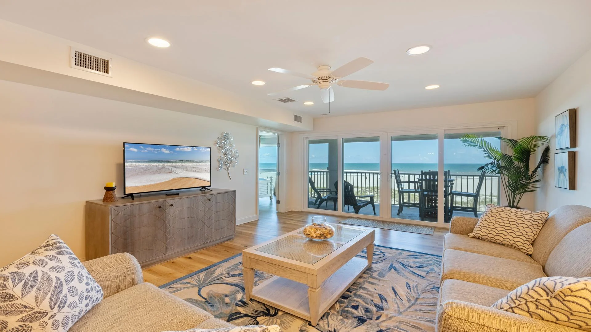 Living room with beige sofas, patterned pillows, wood coffee table on floral rug, and ocean views through sliding glass doors.