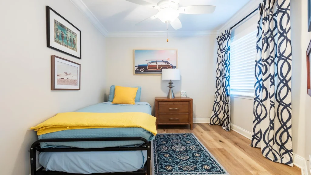 Light wood flooring frames a twin bed with blue and yellow bedding, beside patterned navy-and-white curtains.