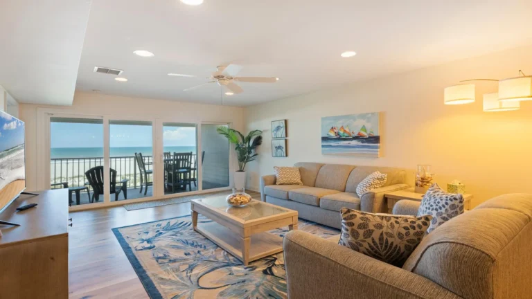 Living room with two beige sofas, patterned pillows, coastal artwork, and sliding glass doors opening to oceanfront balcony.