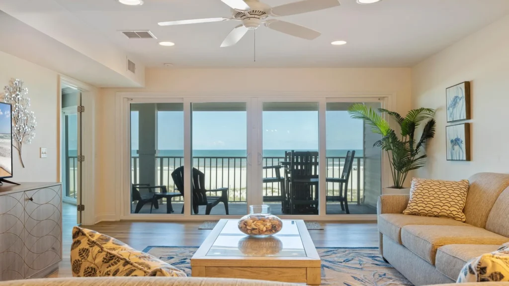 Living room with beige sofas, patterned pillows, glass-top coffee table, and sliding doors opening to ocean-view balcony.