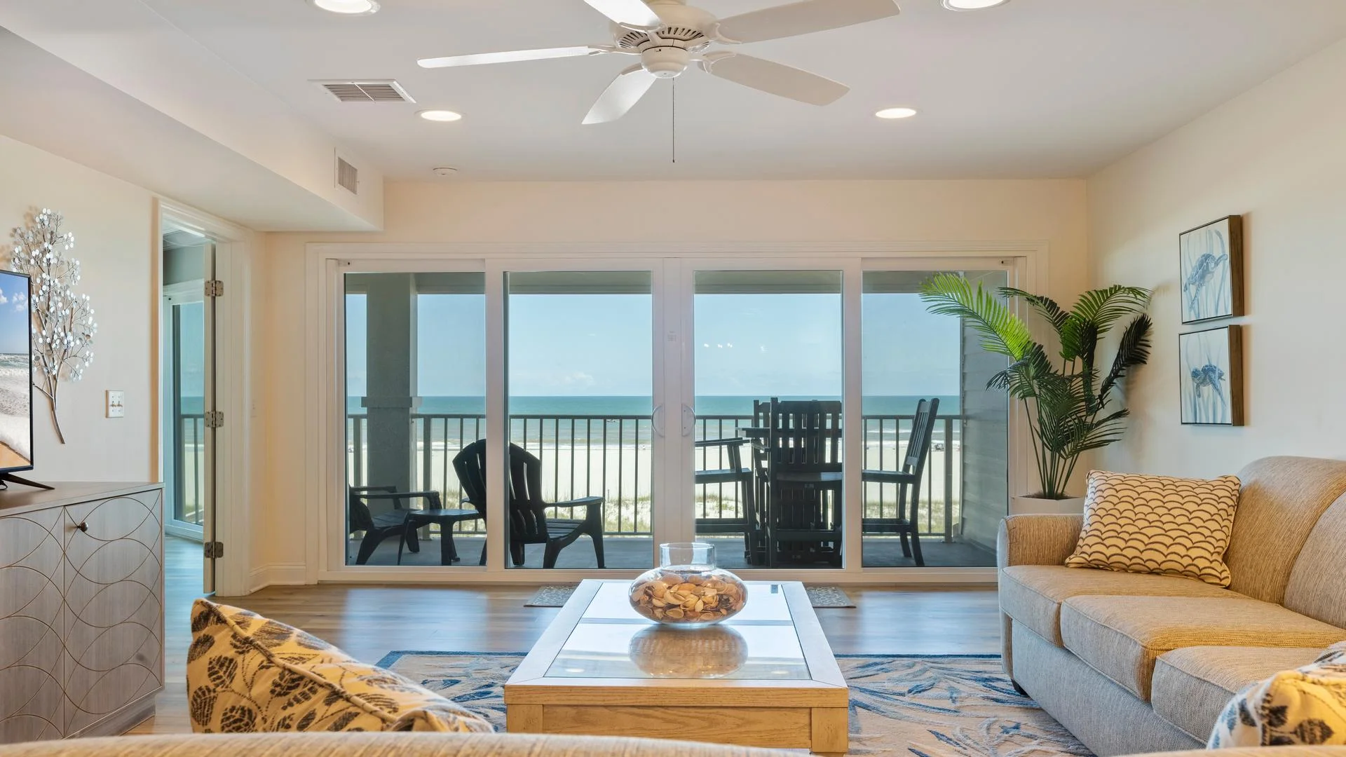Living room with beige sofas, patterned pillows, glass-top coffee table, and sliding doors opening to ocean-view balcony.