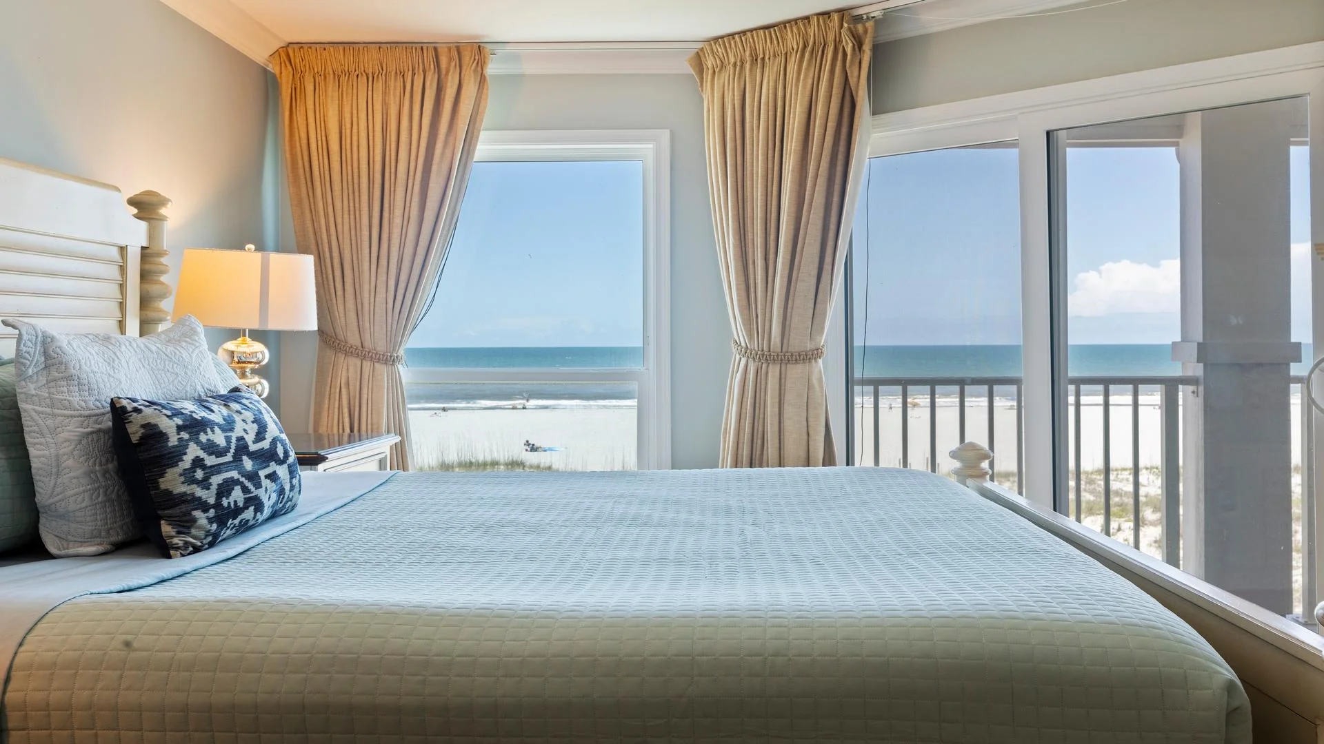 White wooden bedframe with blue quilt and patterned lumbar pillow, flanked by bedside lamp and ocean windows.