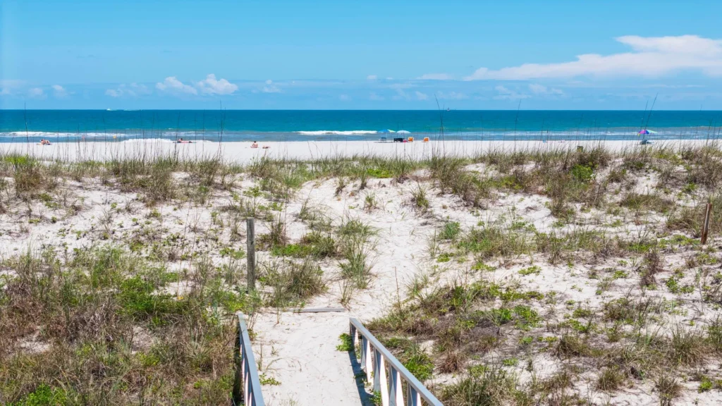 Sandy path bordered by dune grass leads to a white sand beach with Atlantic Ocean waves under a clear blue sky.