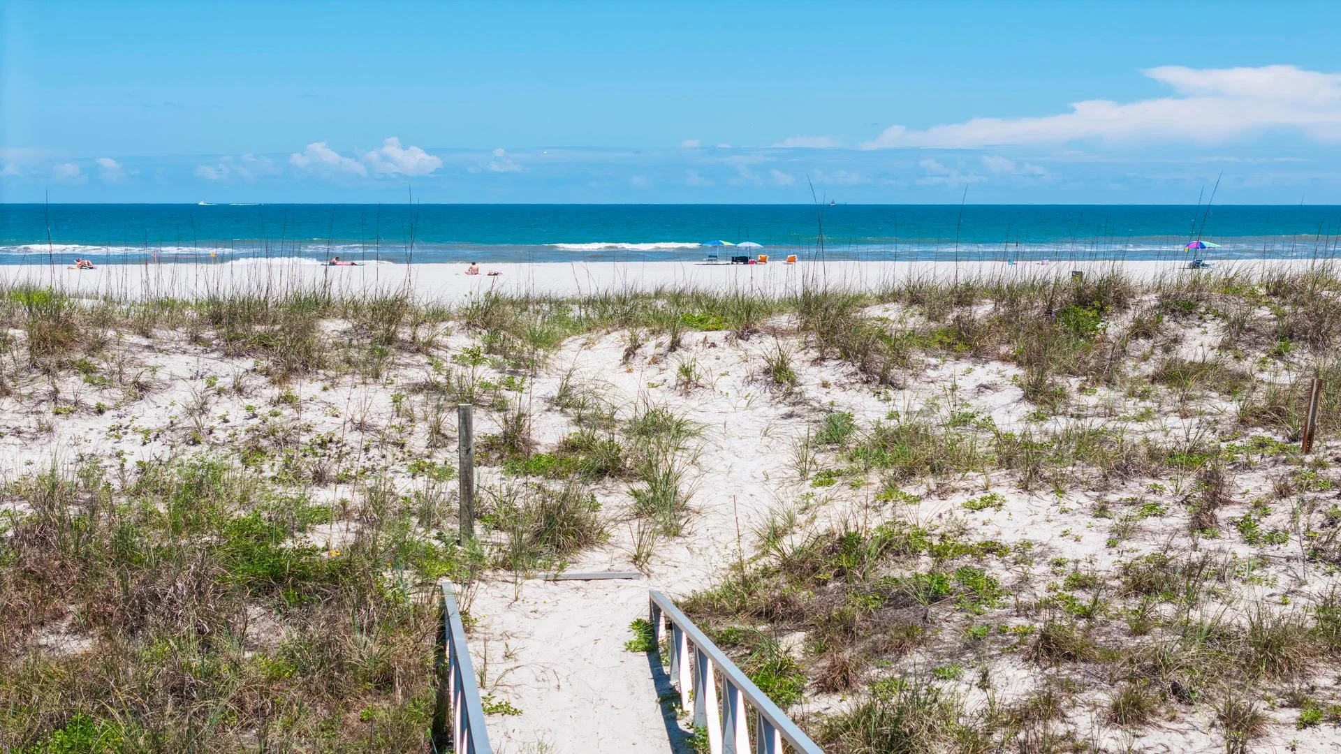 Sandy path bordered by dune grass leads to a white sand beach with Atlantic Ocean waves under a clear blue sky.