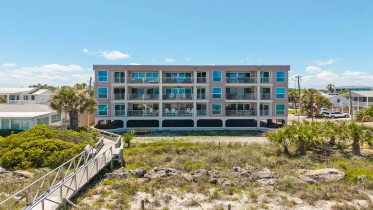 Exterior view of a three-story condo building with balconies overlooking sandy dunes and a wooden boardwalk to the beach.