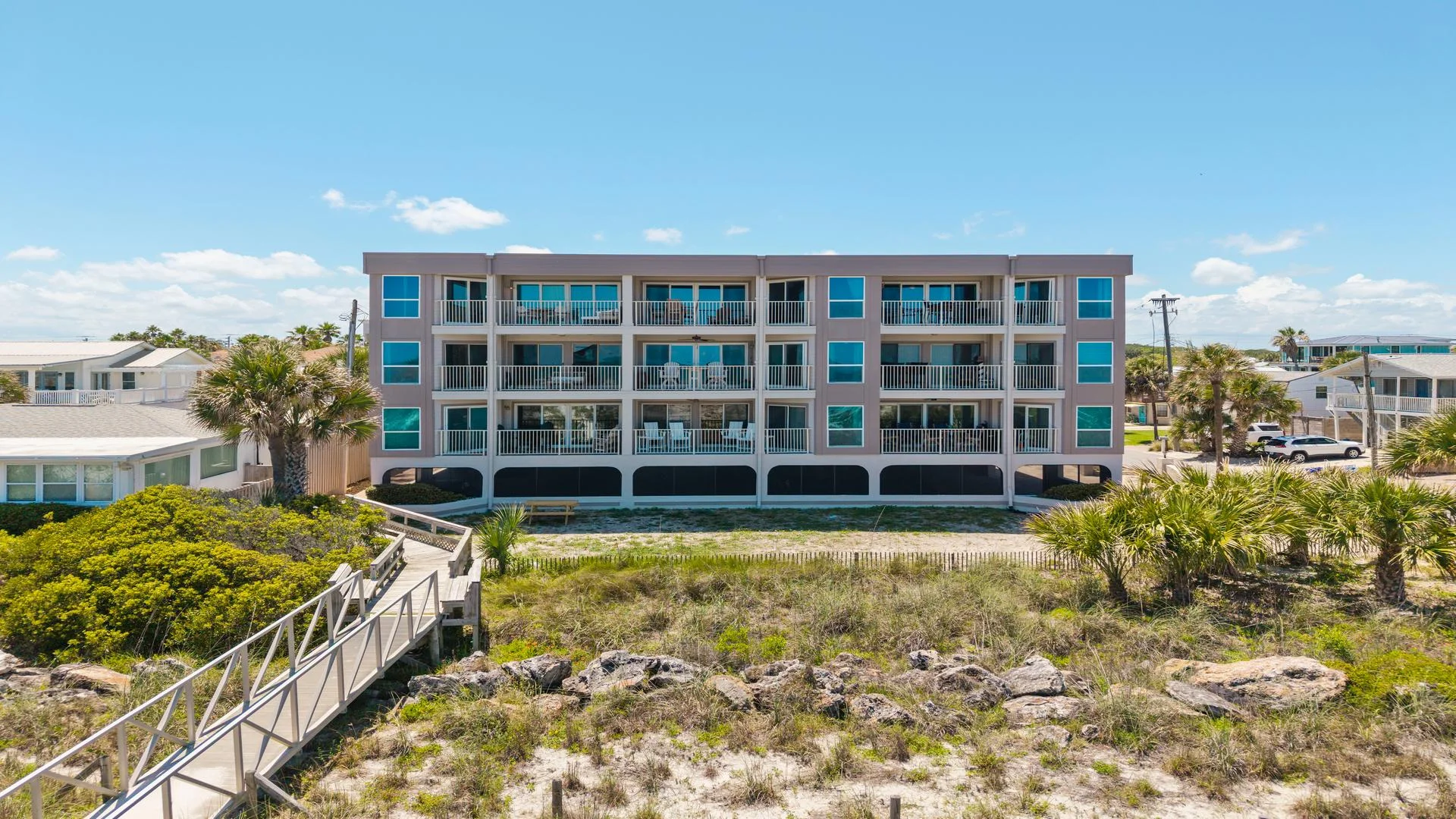 Exterior view of a three-story condo building with balconies overlooking sandy dunes and a wooden boardwalk to the beach.