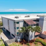 Three-story condo building with light stucco walls, red tile roof accents, and palm trees by a sandy beach.