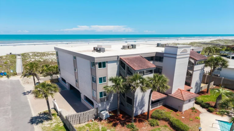 Three-story condo building with light stucco walls, red tile roof accents, and palm trees by a sandy beach.