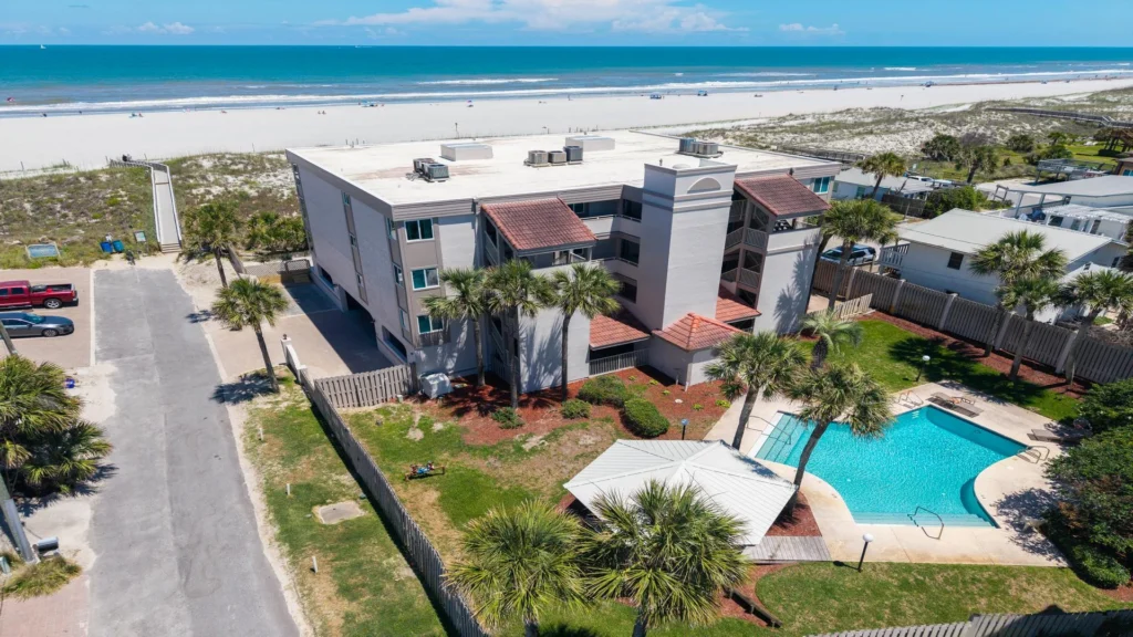 Three-story condo building with red tile roofs, fenced pool area, palm trees, and Atlantic Ocean beach access.
