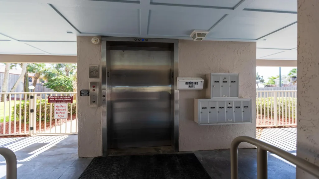Elevator with stainless steel doors next to a wall-mounted mailbox unit, textured beige walls, and gated outdoor area.