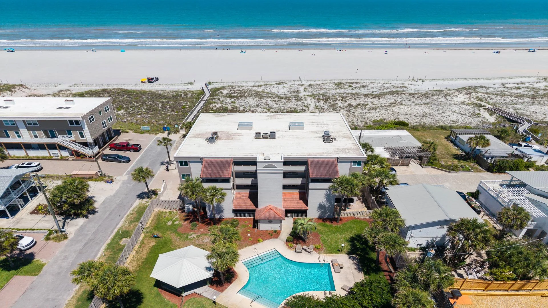 Aerial view of a three-story condo building with a shaped swimming pool, palm trees, and direct beach access.