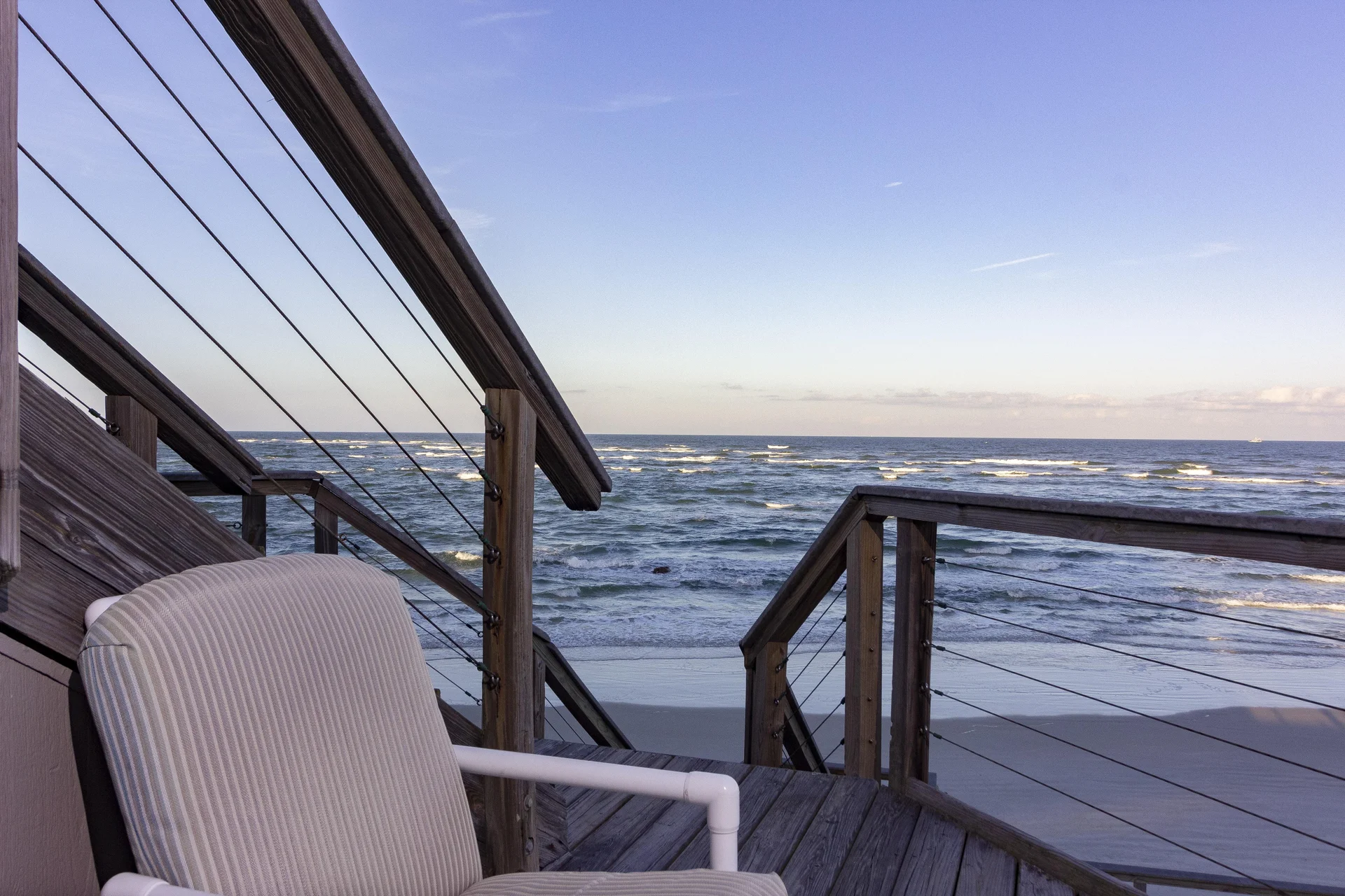 Striped cushioned chair sits on a wooden balcony with cable railings overlooking waves and sandy beach.