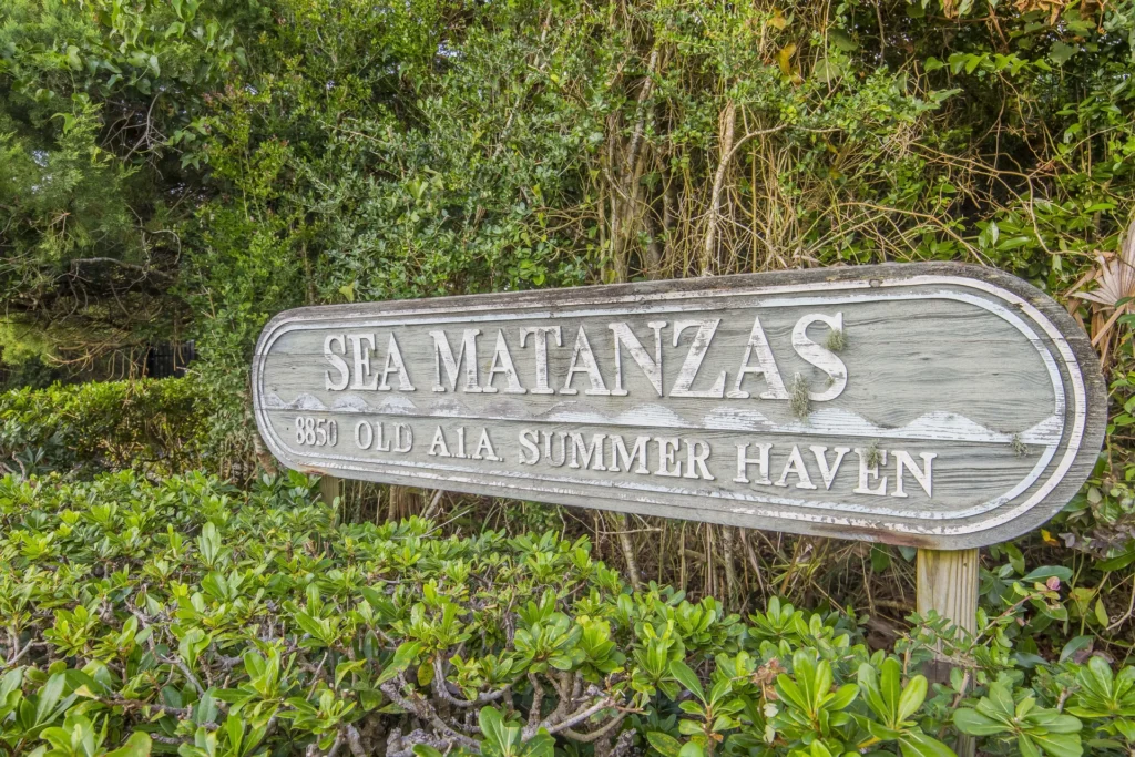 Weathered wooden sign reading "Sea Matanzas 8850 Old A1A Summer Haven" surrounded by green shrubs and trees.