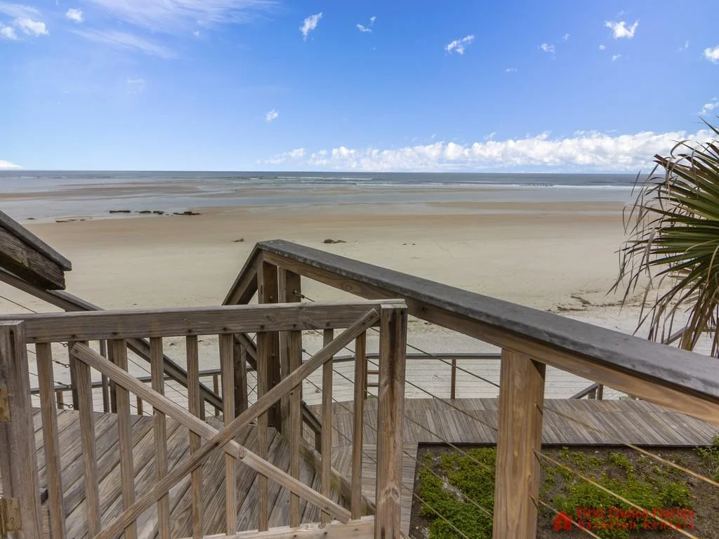 Wooden stair railings lead down to the wide sandy beach under a bright blue sky with scattered clouds.