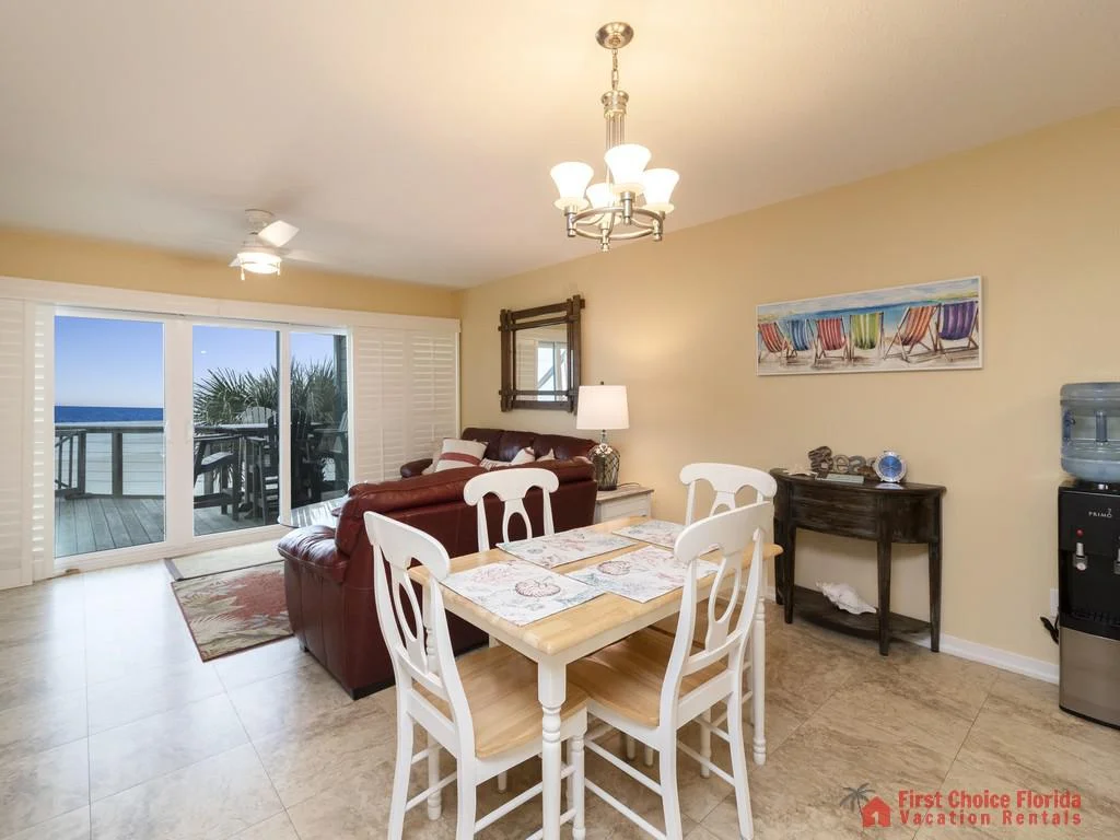 Dining area with a light wood table and white chairs opens to a living room featuring maroon leather sofas and ocean views.
