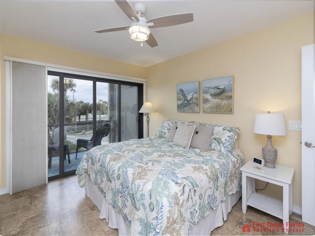 King bed with sea-themed quilt faces sliding glass doors to a balcony overlooking palm trees and a street.