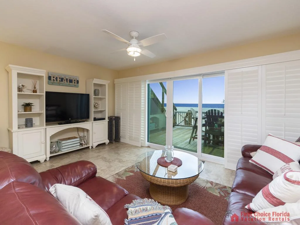 Maroon leather sofas frame a glass-top wicker coffee table with ocean views through sliding patio doors.