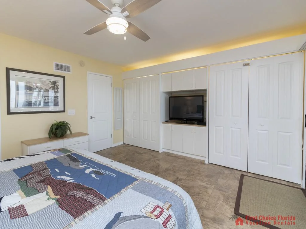Ceiling fan with light fixture turns above a bed dressed in coastal-themed quilt and a dresser topped with a potted plant.
